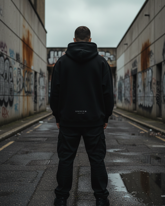 A man wearing a black Craftklart Authority heavyweight oversized hoodie with drop shoulders in an urban alleyway.